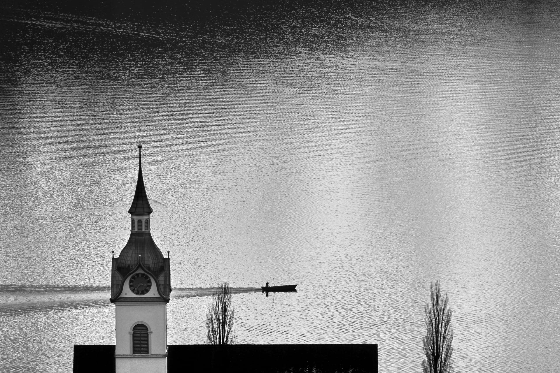 Kirchturm am Zugersee, dahinter ein Fischerboot auf dem gleissenden Wasser